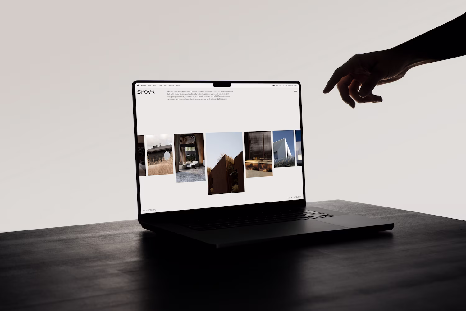 Laptop on a dark wooden table displaying an architecture portfolio website with a hand reaching toward the screen from the right.