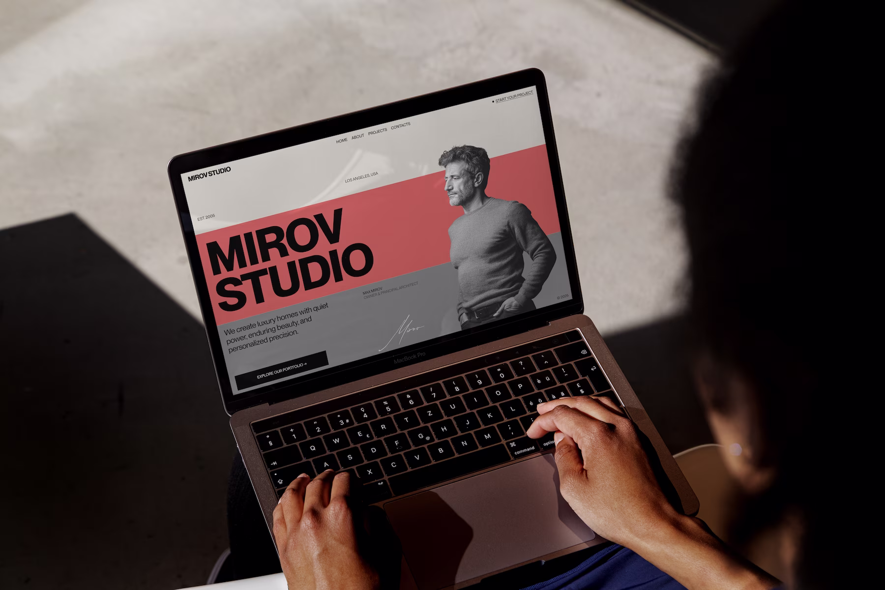 Person typing on a MacBook Pro showing a website for Mirov Studio featuring a grayscale photo of a man, text, and navigation links.
